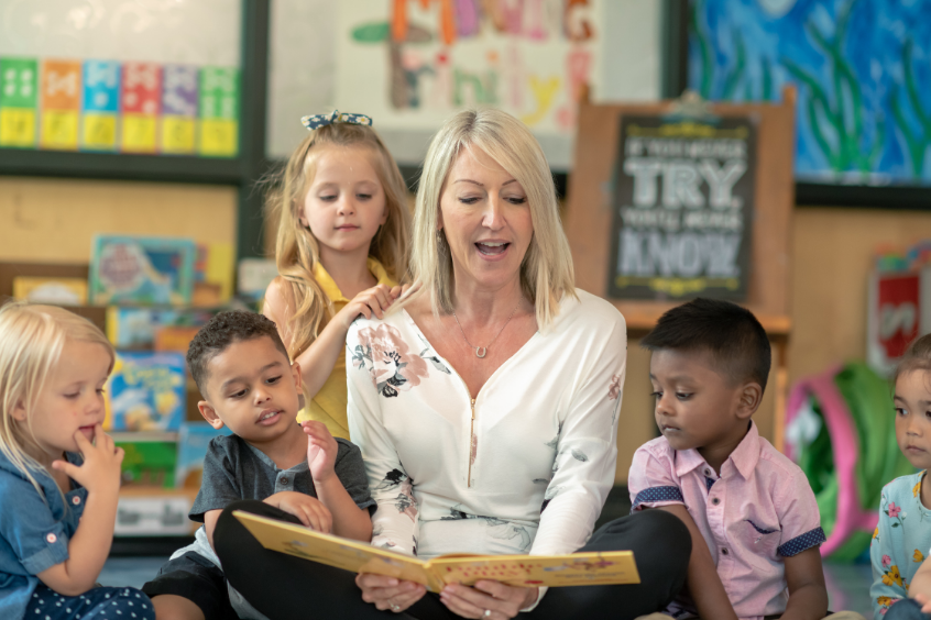 A preschool teacher sitting on the floor reading a book to her students who are huddled around her listening and smiling