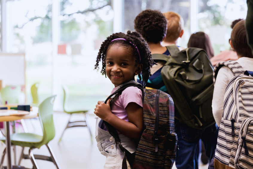 Adorable little girl smiles in her kindergarten classroom wearing a backpack